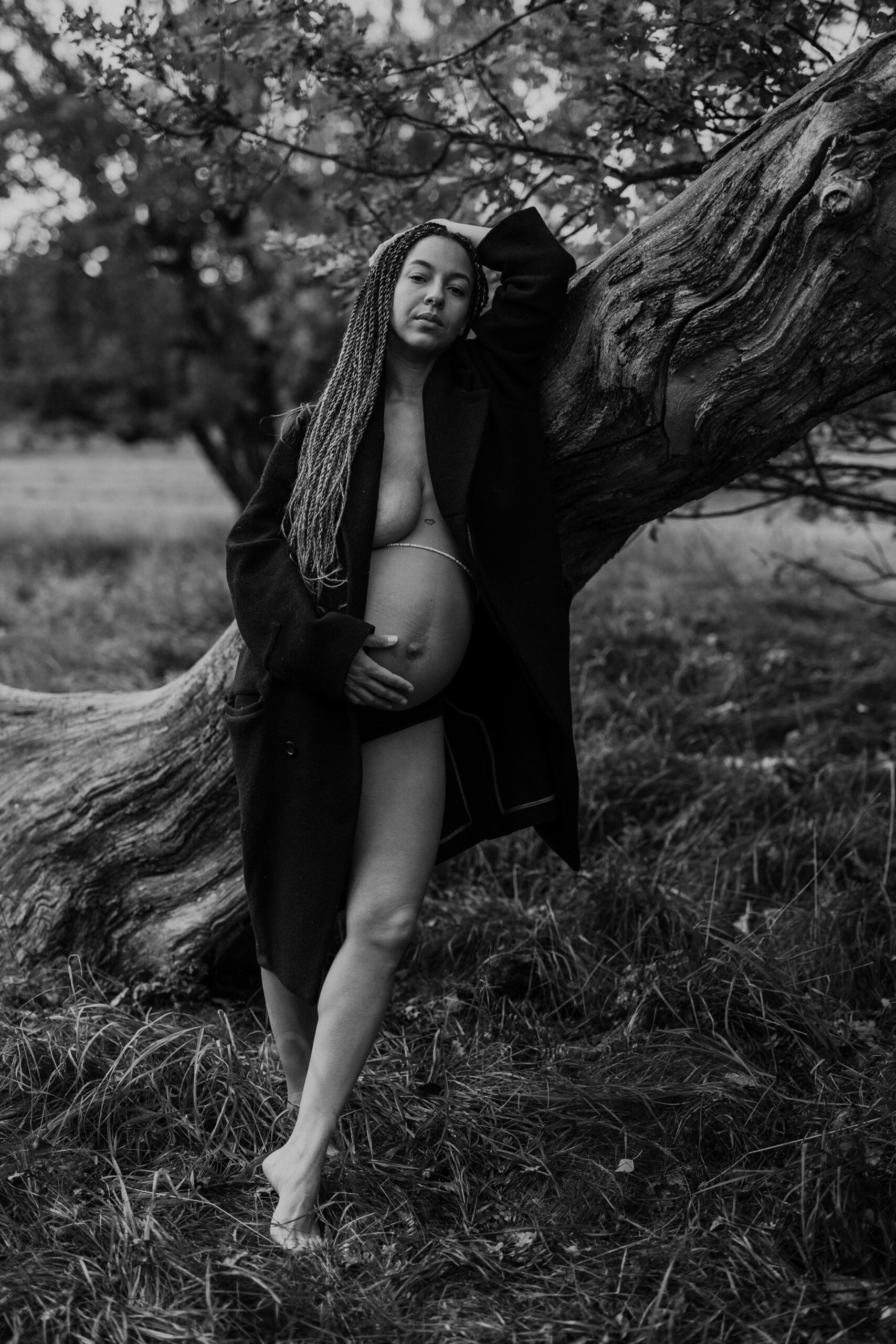 gravid kvinna står utomhus i svart kappa och barfota i gräset lutad mot en stock
