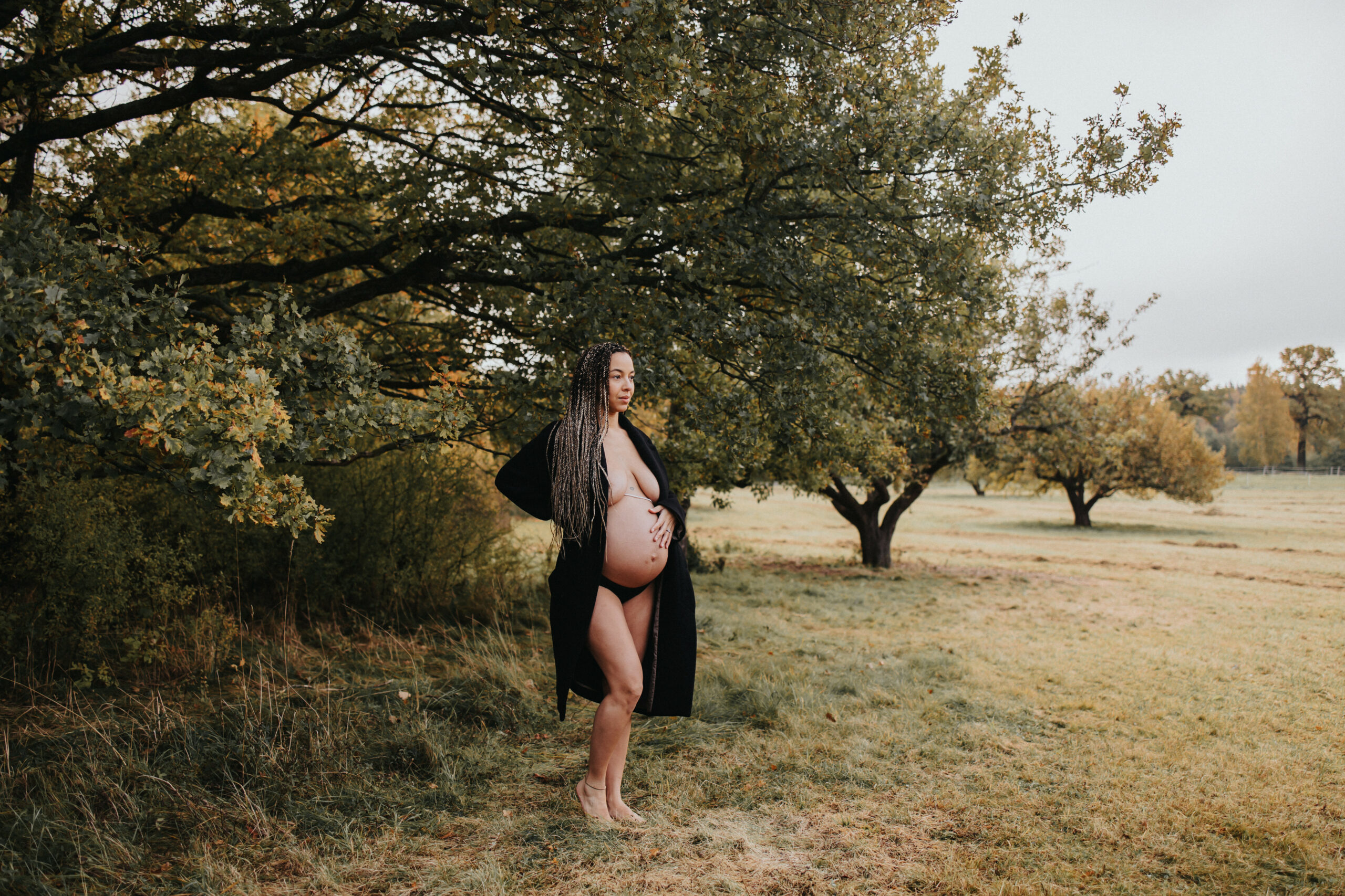 gravid kvinna står utomhus i svart kappa och barfota i gräset under en stor ek