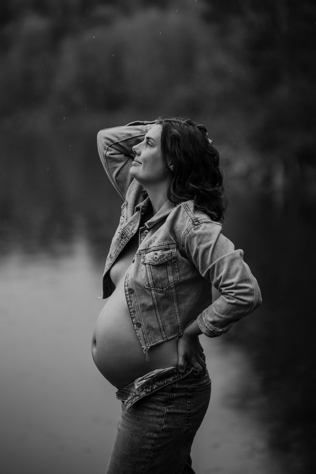 gravid kvinna står i regnet med ansiktet mot himlen och har jeans och jeansjacka på sig