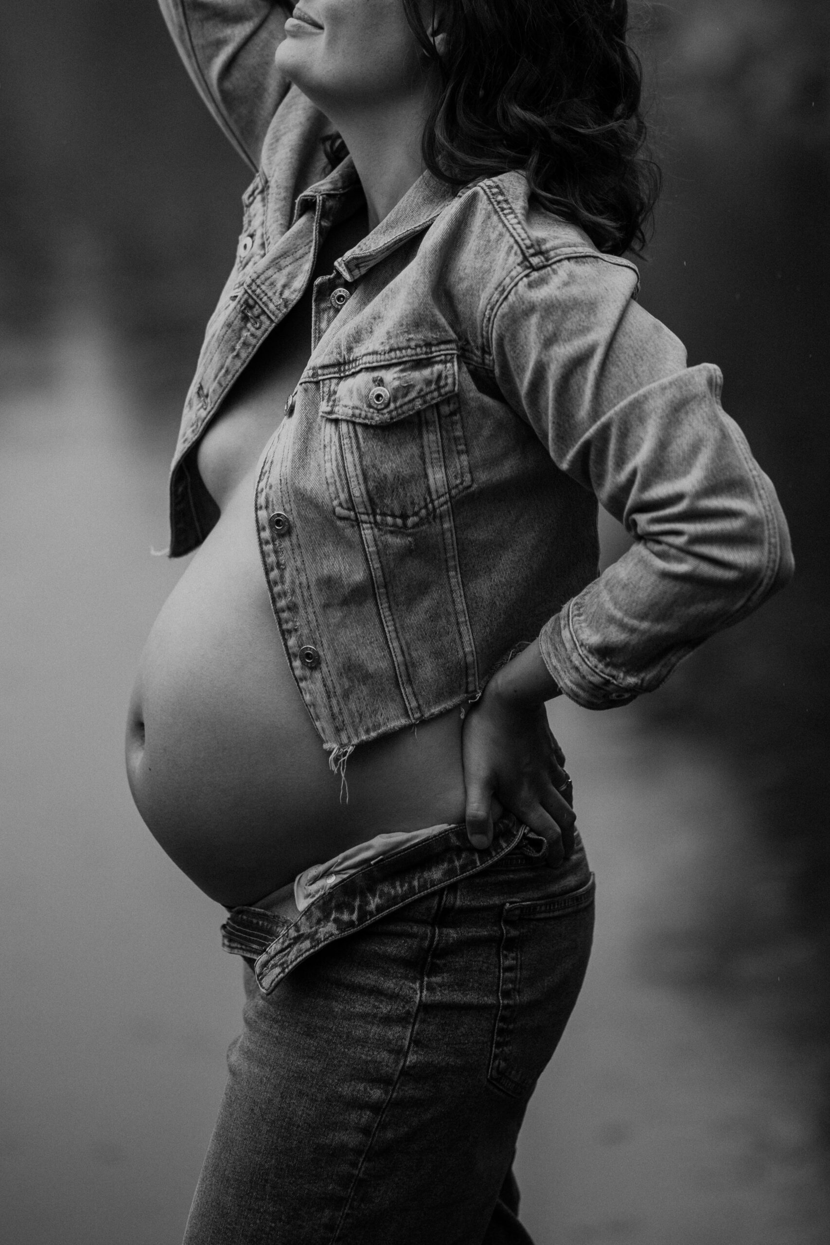 Gravidfotografering i regnet – svartvit bild av gravid kvinna som står i regnet med öppen jeansjacka