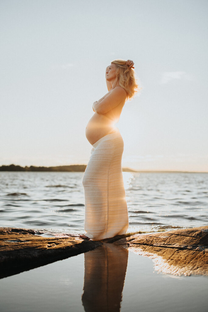 gravid kvinna står i vit kjol i solnedgången vid vattnet